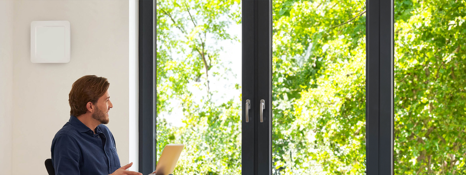 Moderne Wohnung mit Wandlüfter und großem Fenster, durch das grüne Bäume sichtbar sind.